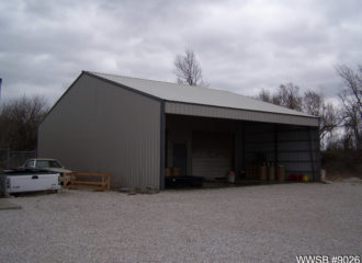 A metal storage shed with an open front reveals various items inside, hinting at the versatility of metal building homes. It sits on a gravel surface under overcast skies, with bare trees silhouetted in the background and a white vehicle partially visible to the left.