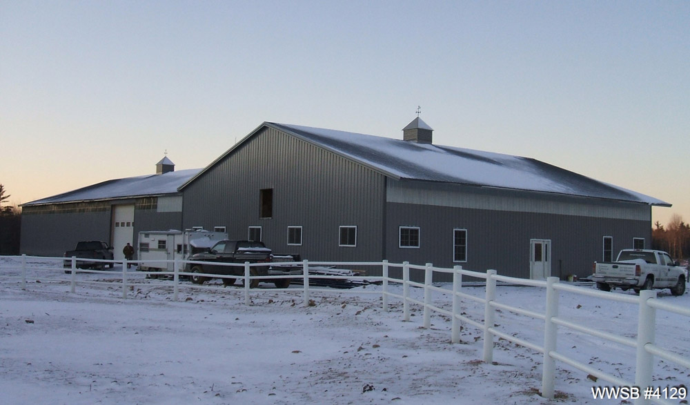 A large, gray barn with a snow-covered roof sits behind a white fence. Nearby, several vehicles and equipment rest under clear skies suggesting a cold winter day. Adjacent to the barn, metal garage kits offer seamless storage for an array of tools and machinery.