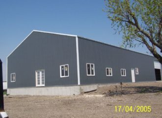 A large, gray, metal warehouse building with a slanted roof on a concrete foundation. It has multiple windows and a white door. The ground around it is gravelly, with a leafless tree on the right. The photo date is April 17, 2005.