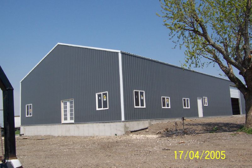 A large, gray, metal warehouse building with a slanted roof on a concrete foundation. It has multiple windows and a white door. The ground around it is gravelly, with a leafless tree on the right. The photo date is April 17, 2005.