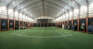 The indoor tennis court features green flooring and white lines, housed within a spacious building with high walls. Transparent ceiling panels allow natural light to flood the space, while tennis equipment is neatly scattered along the sides.