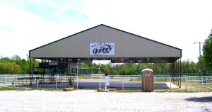 A large open pavilion with a metal roof labeled "Miller Pavilion Lite" stands in a gravel area, reminiscent of a fair building. A portable toilet is on the right side, and county fair-style picnic tables are underneath. Trees and greenery are visible in the background.