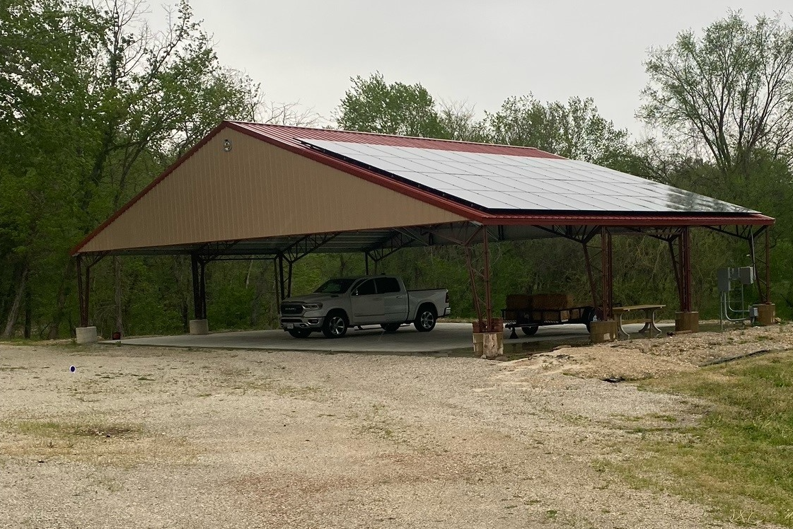 In the tranquil setting of Cedar Hollow, an agricultural metal pavilion with a red roof and solar panels shelters a pickup truck, trailer, and picnic table. The structure rests on a gravel surface, encircled by lush trees.