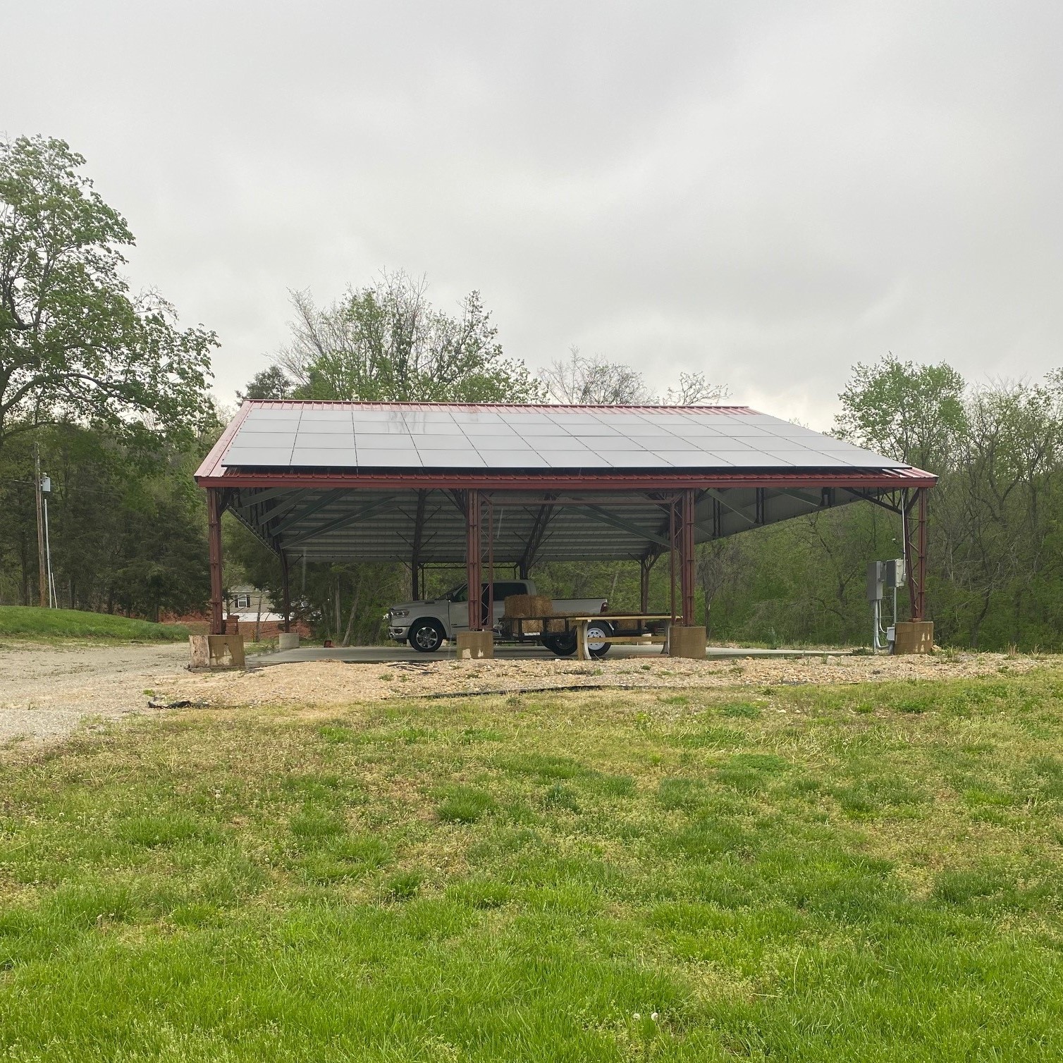 A metal carport with a solar panel roof stands in Cedar Hollow's lush greenery. This agricultural-style pavilion shelters two vehicles, surrounded by trees under the cloudy skies.