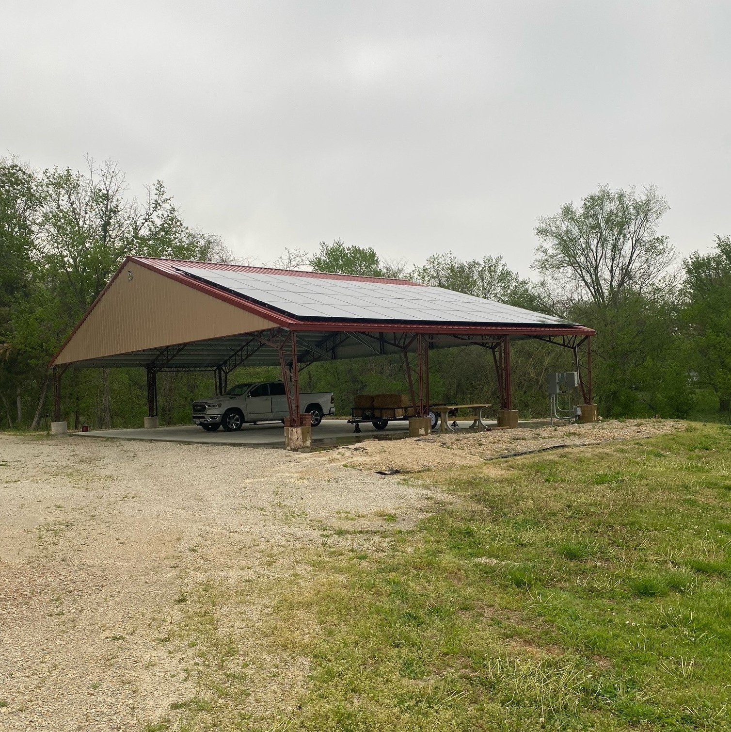 Nestled in Cedar Hollow, this agricultural pavilion features an open-sided, metal-roofed design. A pickup truck and trailer are parked beneath it, while a picnic table and chairs invite gatherings. Surrounded by trees under a cloudy sky, it's a serene retreat in the heart of nature.