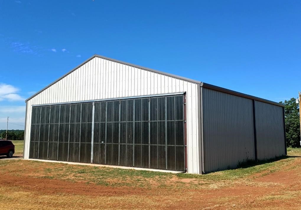 A large white metal barn, reminiscent of hangar design, with closed black sliding doors stands on a dirt and grass lot under a clear blue sky.