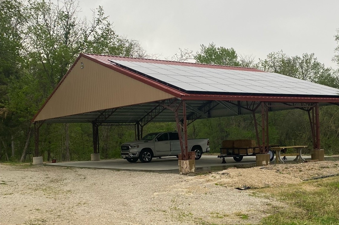 A metal carport, reminiscent of the Cedar Hollow Agricultural Pavilion, boasts solar panels on its roof, sheltering a white pickup truck and construction materials. Surrounded by trees and grass, it creates a serene rural setting.