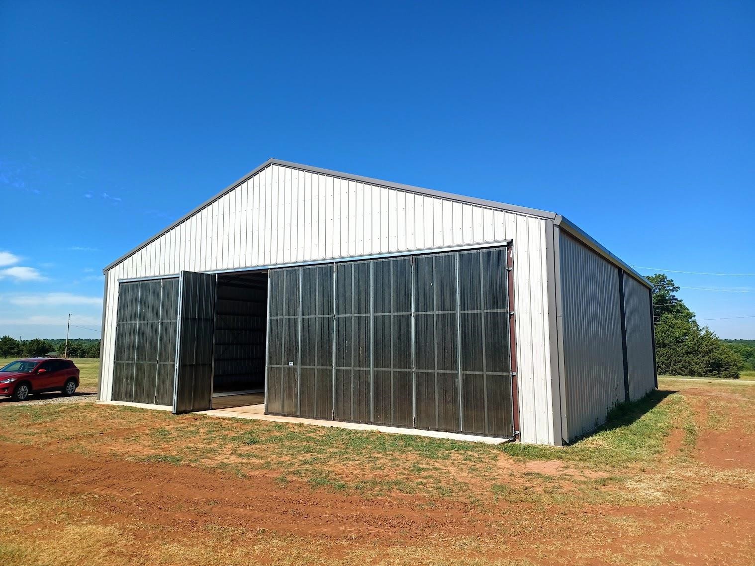 A metal barn, echoing a hangar design with large open sliding doors, stands on a grassy field under a clear blue sky. A red car is parked nearby on the left.