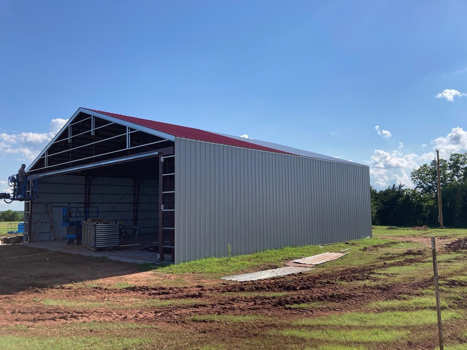 A large metal structure, echoing an aircraft hangar design, stands with a partially open front in a grassy area. Its red roof gleams under the clear sky dotted with a few clouds, while construction materials are scattered inside, hinting at the park hangar's ongoing development.