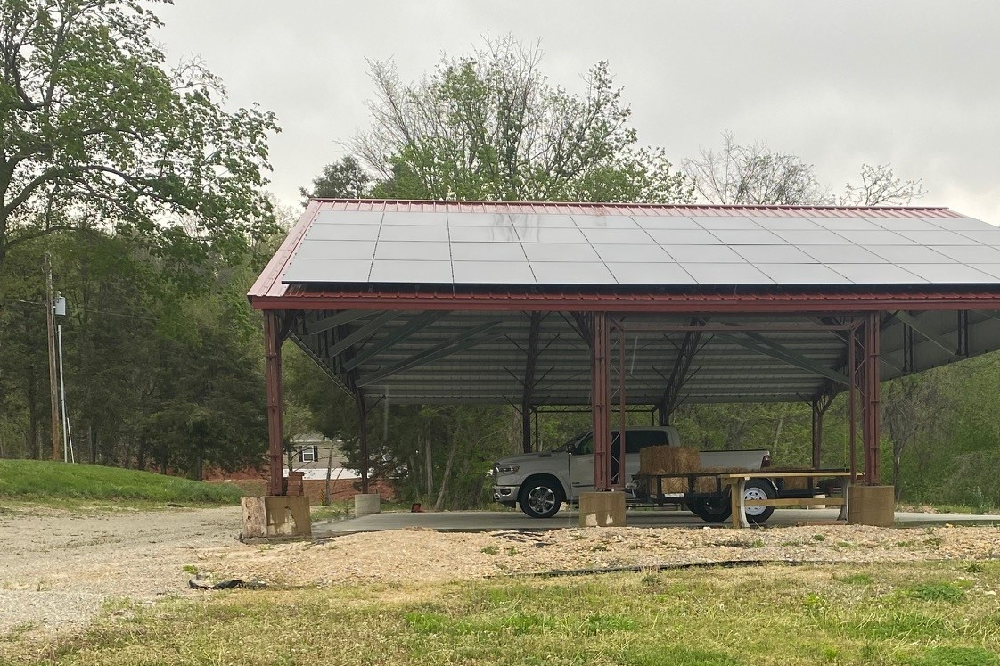 A solar-paneled carport, resembling an Agricultural Pavilion, shelters a gray pickup truck with a flatbed trailer nearby, while Cedar Hollow's lush trees and grass frame the scene under an overcast sky.