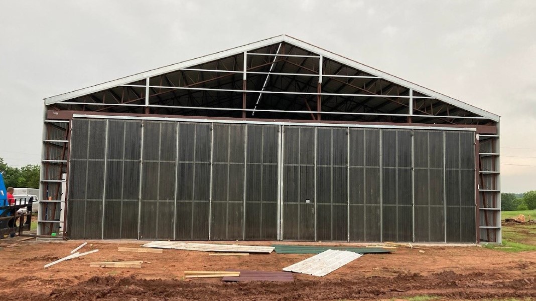 A large, partially constructed metal structure stands on a muddy construction site, its design reminiscent of a hangar. The building features a high, triangular roof and large sliding doors. Overcast skies suggest recent or impending rain as scattered building materials hint at its future as an aircraft haven.