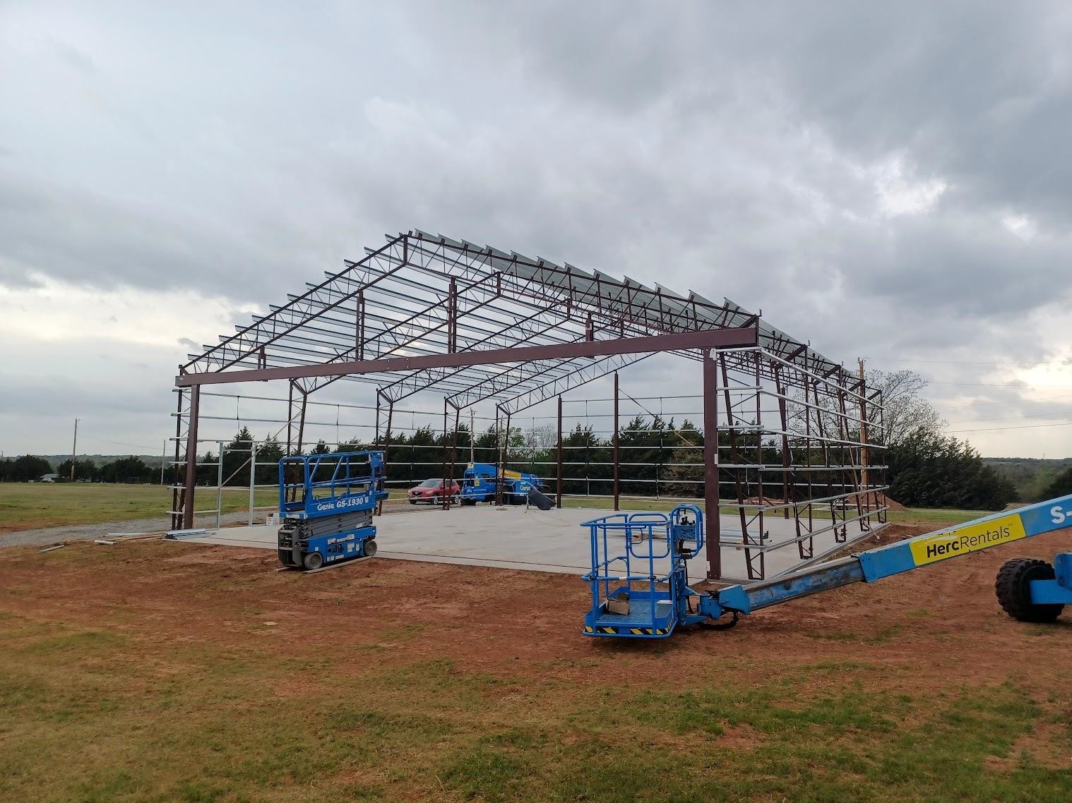 A steel frame of a large aircraft hangar under construction on a concrete foundation. Two blue boom lifts are positioned nearby. The scene is set in an open field with cloudy skies above, showcasing the meticulous hangar design amidst sprawling landscapes.