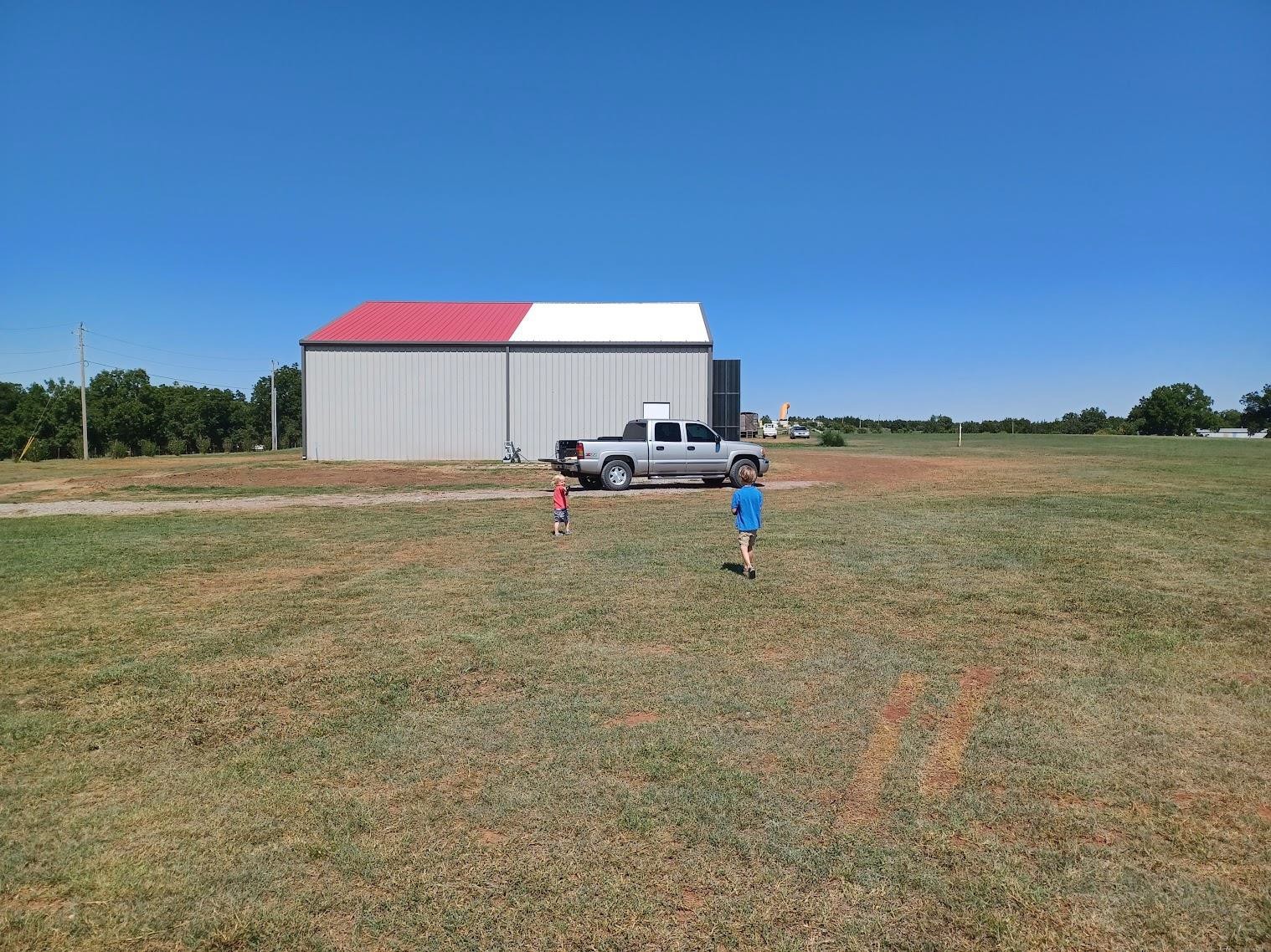 Two children walk toward a gray building with a red roof, echoing hangar design, on a grassy field under a clear blue sky. A silver pickup truck is parked beside the building, resembling an aircraft ready for takeoff.