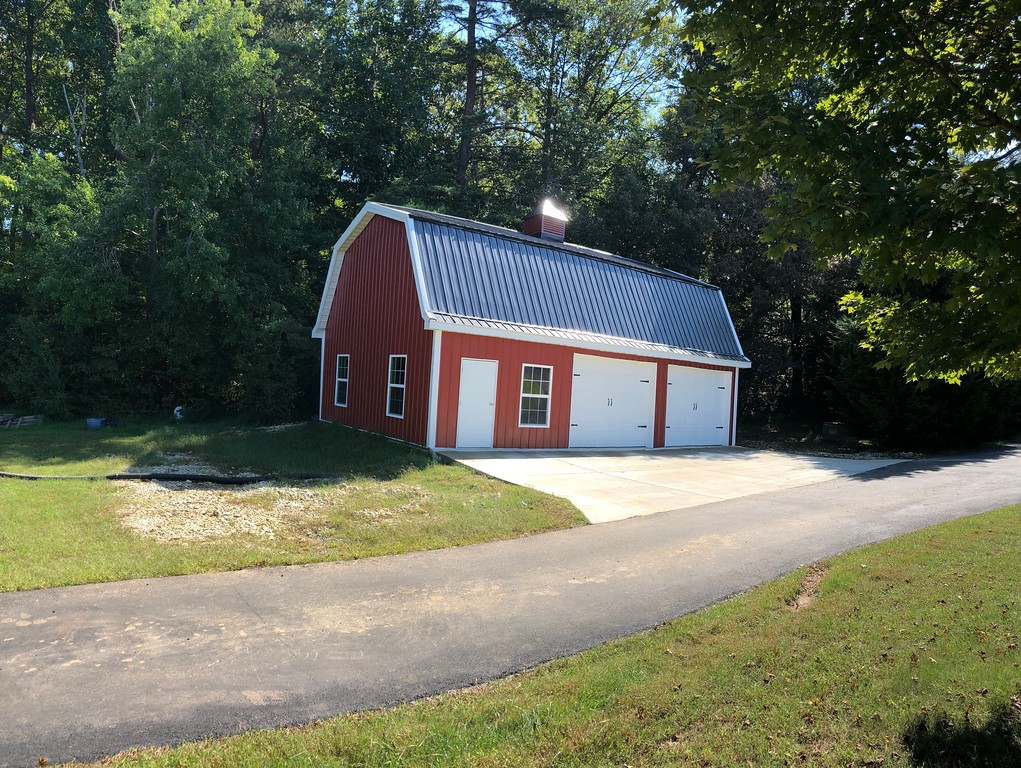 A gambrel-style red barn garage with a black metal roof, white doors, and two windows stands beside a paved driveway. It's surrounded by lush green trees and grass under a clear blue sky.