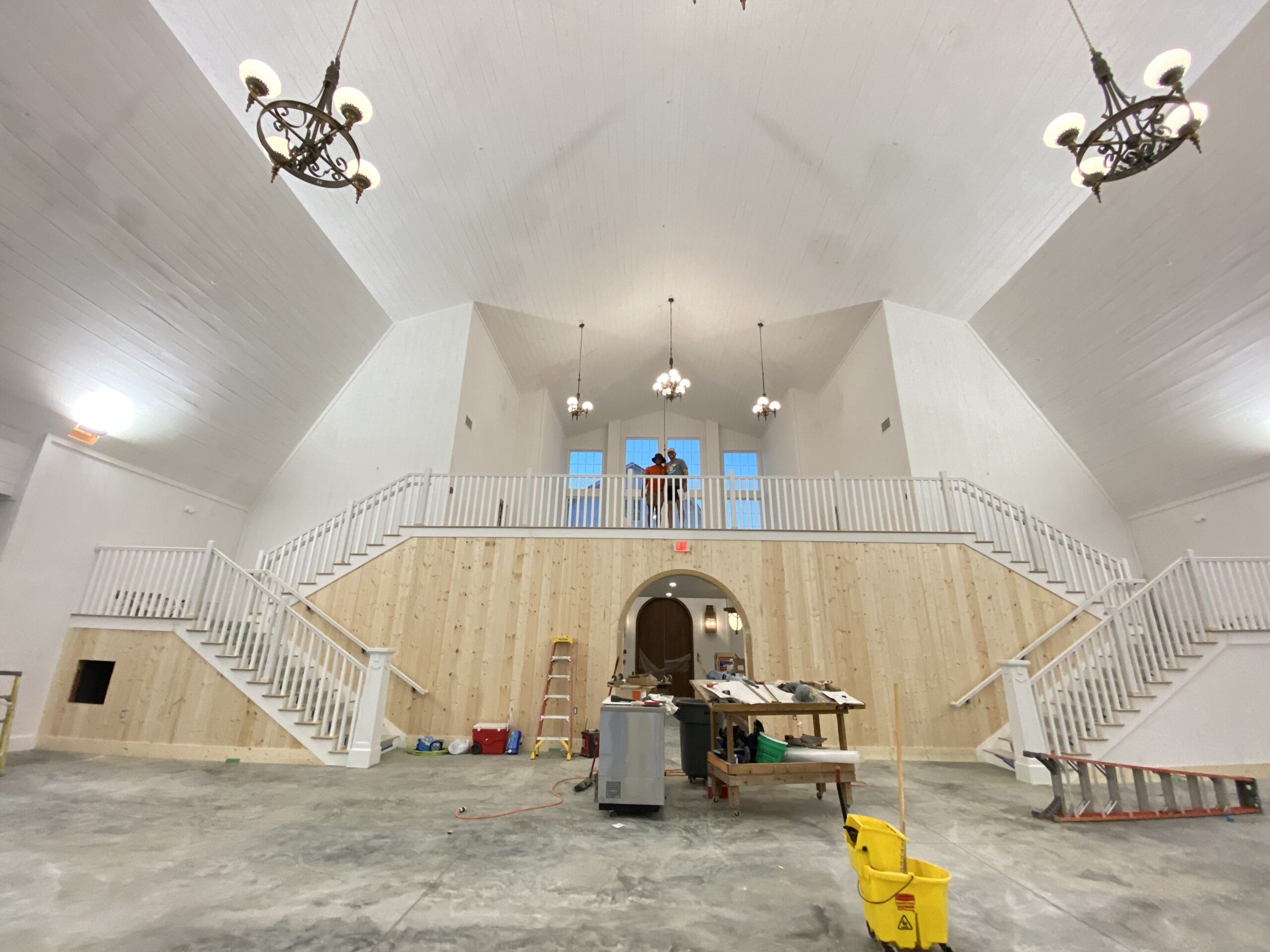 A large, unfinished venue with high ceilings, chandeliers, and wooden walls exudes a charming ambiance. Two elegant staircases lead to a balcony where two people stand envisioning their wedding. Construction materials and tools are scattered across the gambrel-style floor.