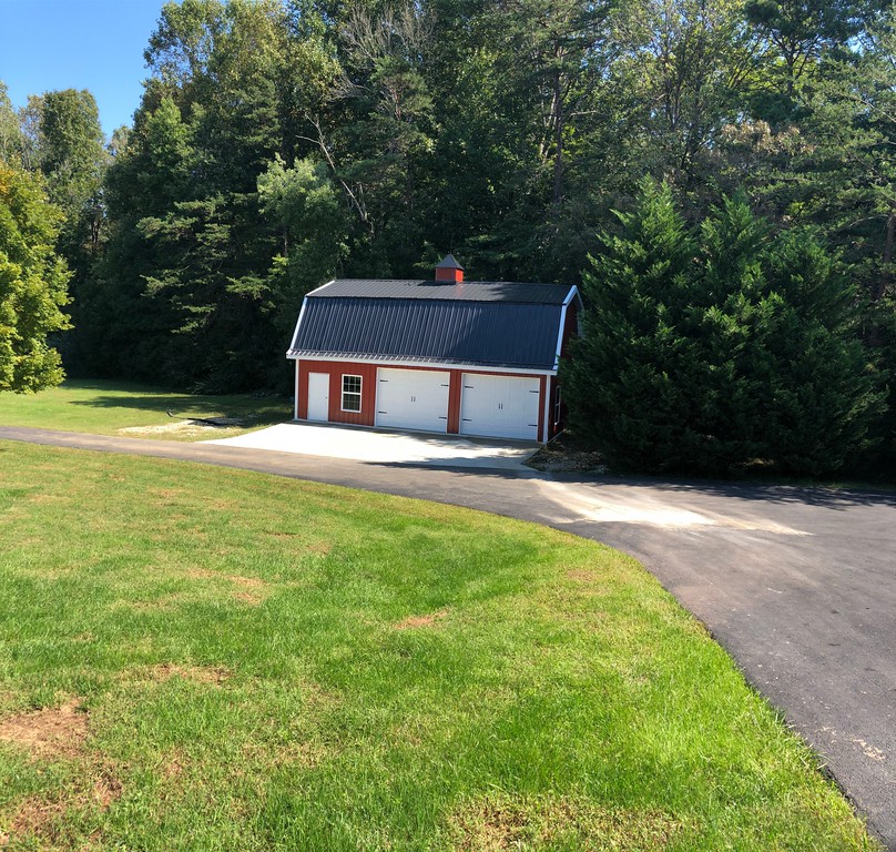 A small, red barn with white trim and a black roof sits at the edge of a paved driveway, resembling a charming countryside shop. It's surrounded by lush green trees and grass on a sunny day.