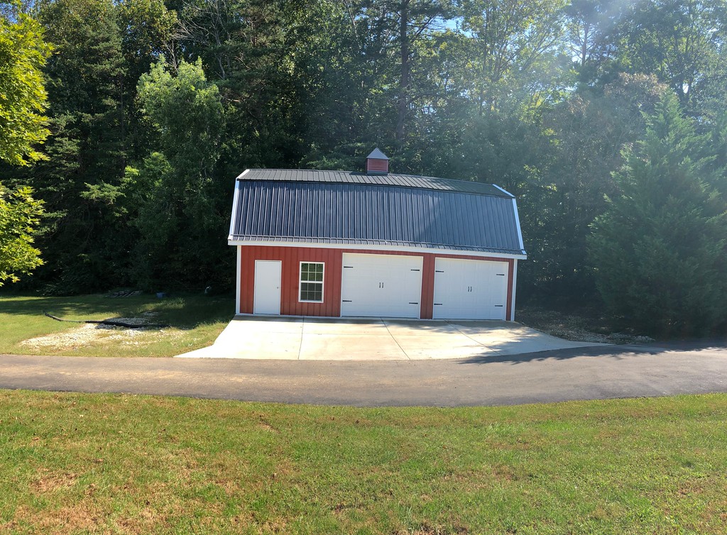 A red barn-style garage with a black metal gambrel roof and two white double doors stands on a concrete driveway. It's surrounded by greenery, including trees and well-kept grass, under a clear blue sky.