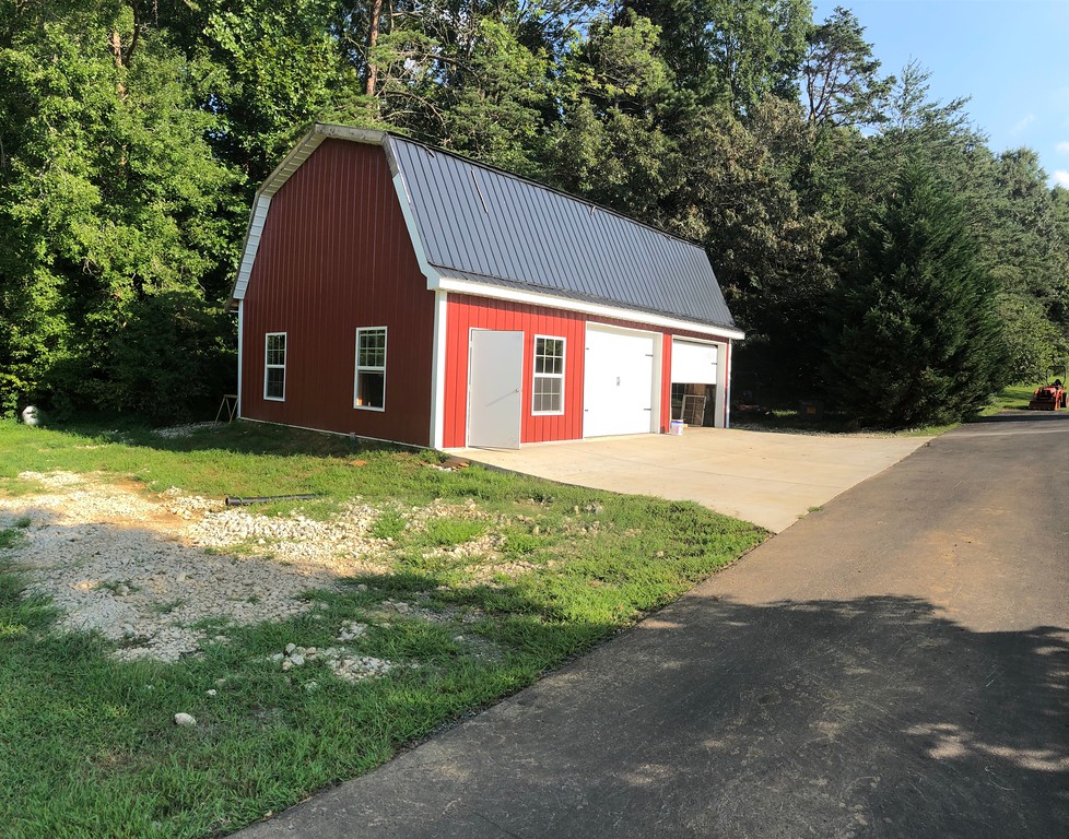 A gambrel-style red barn garage with a white door and metal roof sits adjacent to a driveway. It's surrounded by lush greenery, including trees and grass, under a clear sky.