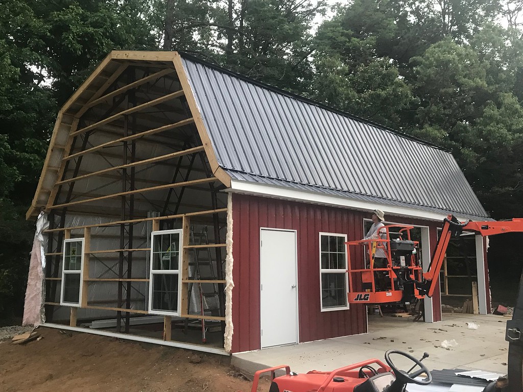 A partially constructed red barn with white trim and a black metal roof, reminiscent of a classic gambrel design, is nestled among trees. A worker on a cherry picker is installing siding on the open front portion of this future shop. A concrete slab lays in the foreground.