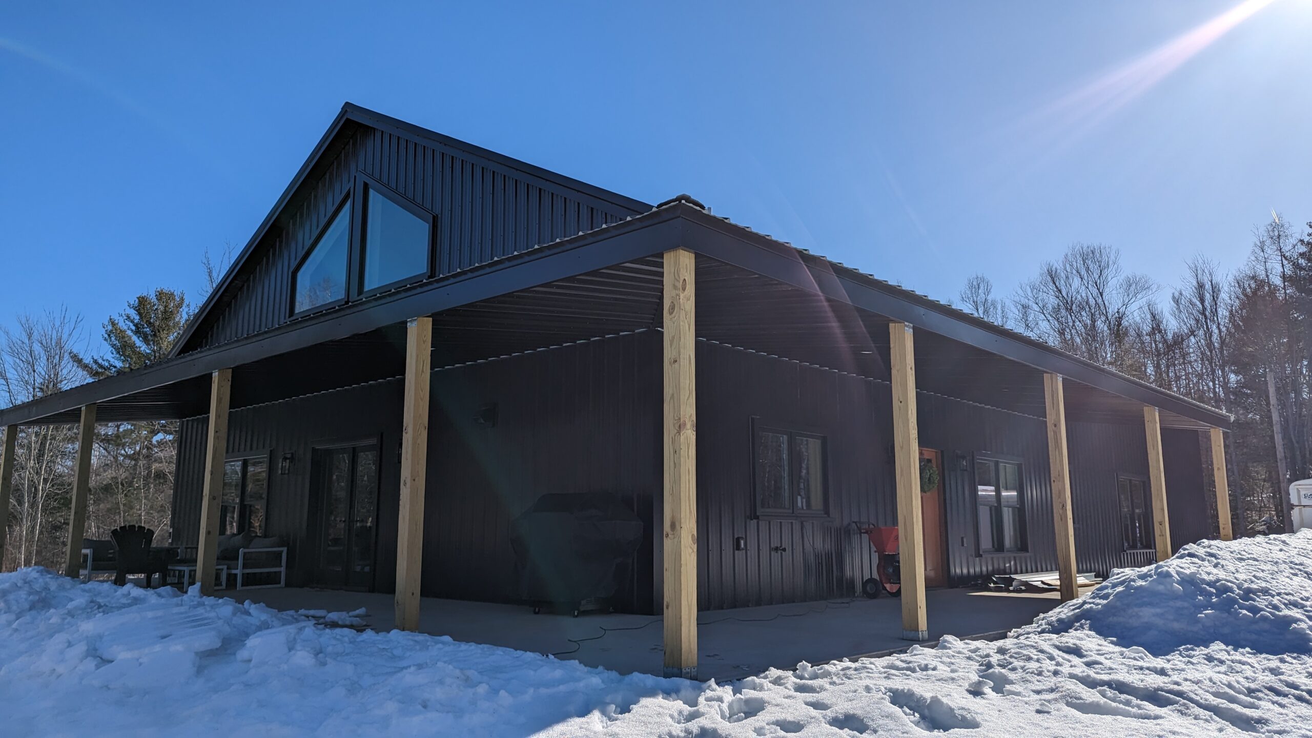 A modern black cabin with a sloped roof and large windows sits in a snowy landscape, reminiscent of a birch tree barndominium. Wooden posts support a wraparound porch while bright sunlight casts shadows on the snow. Sparse trees are visible in the background under a clear blue sky.