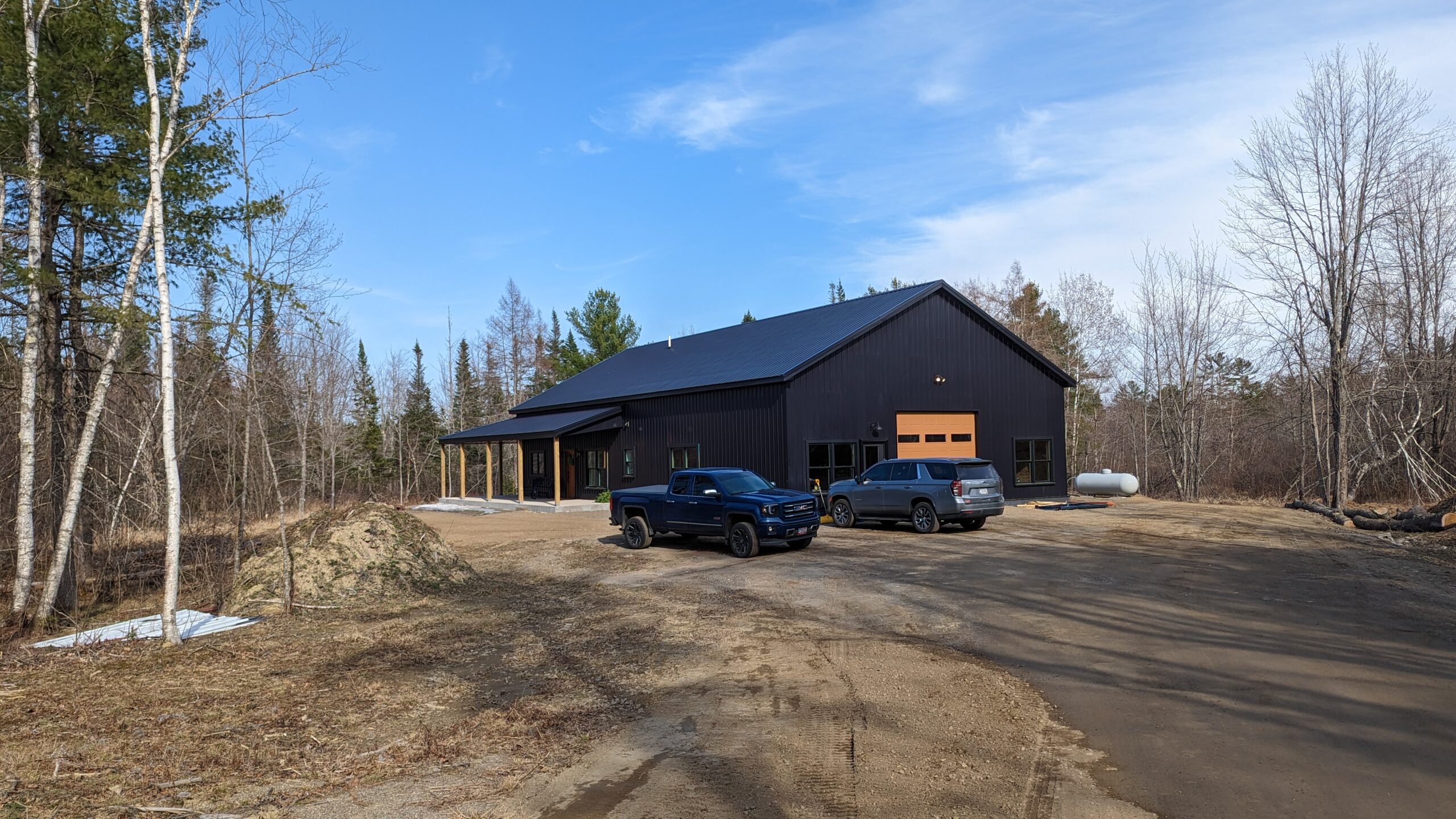 A sleek black barndominium with large windows and a garage stands among trees under a clear blue sky. Two parked vehicles are on the dirt driveway. Leafless birch trees suggest early spring or late autumn, framing this blend of rustic charm and modern design.