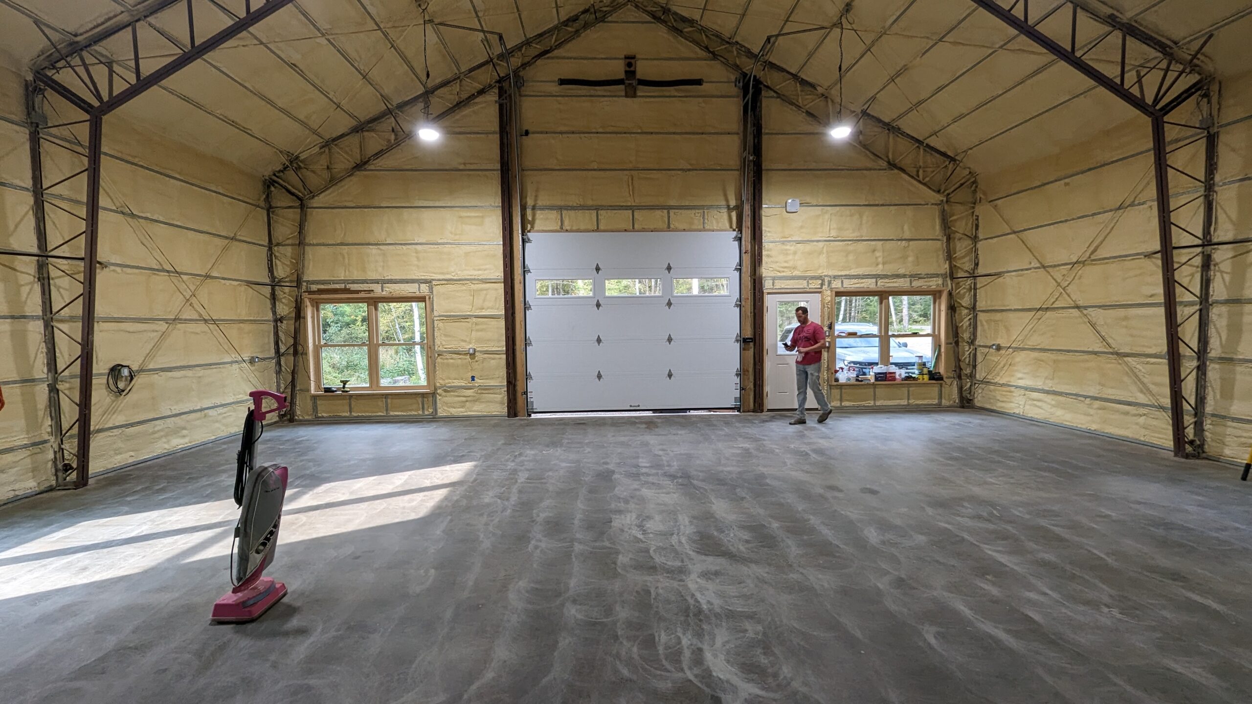 The expansive interior of a large, empty warehouse resembles a modern barndominium with its concrete floors and partially insulated walls. A person stands near the garage door on the right, while a red floor-cleaning machine rests to the left, bathed in natural light filtering through oversized windows.