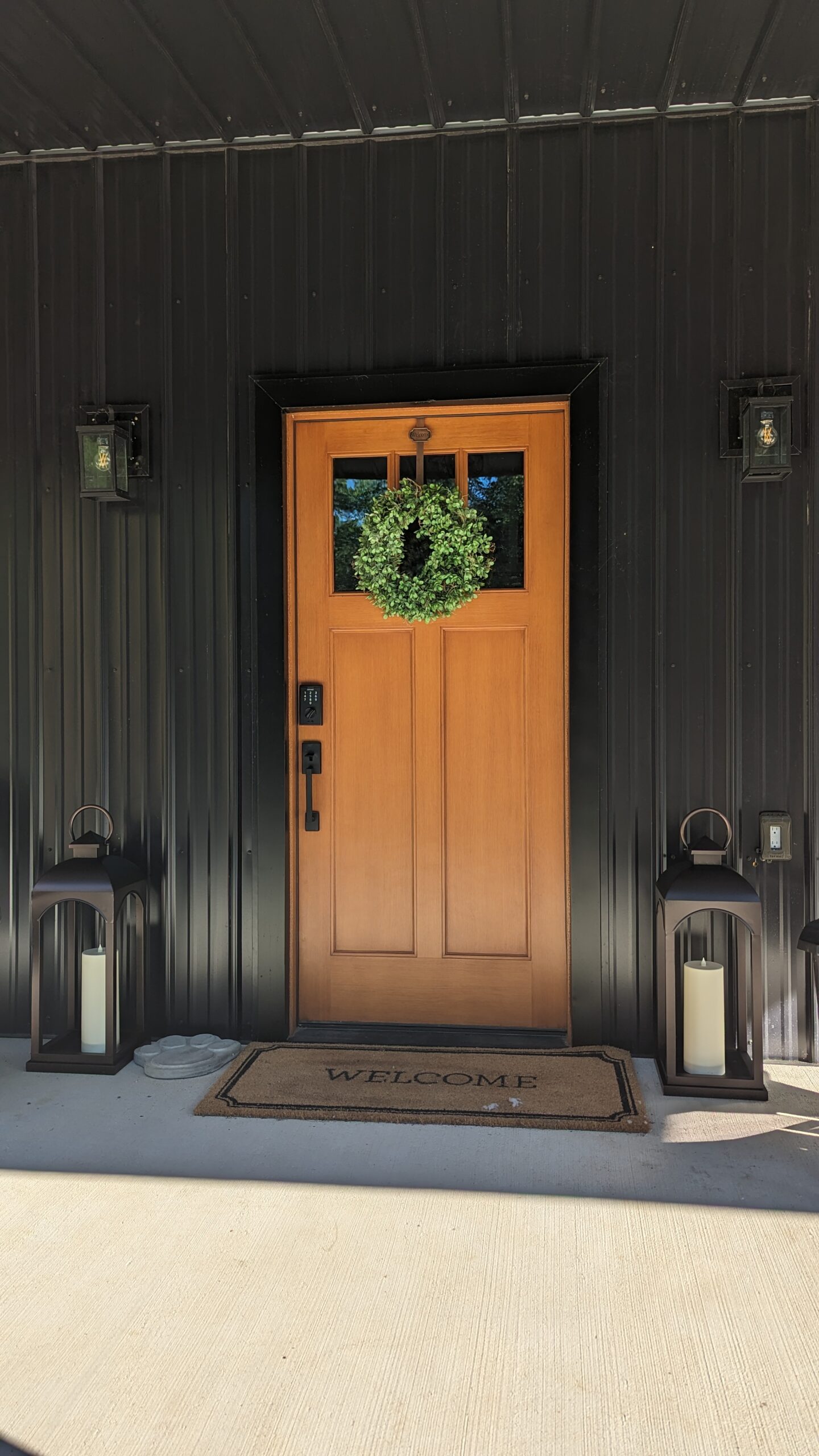 A wooden door with a small window and a green wreath adds charm to the black-walled barndominium. Flanked by birch tree accents, two large lanterns with white candles illuminate the 