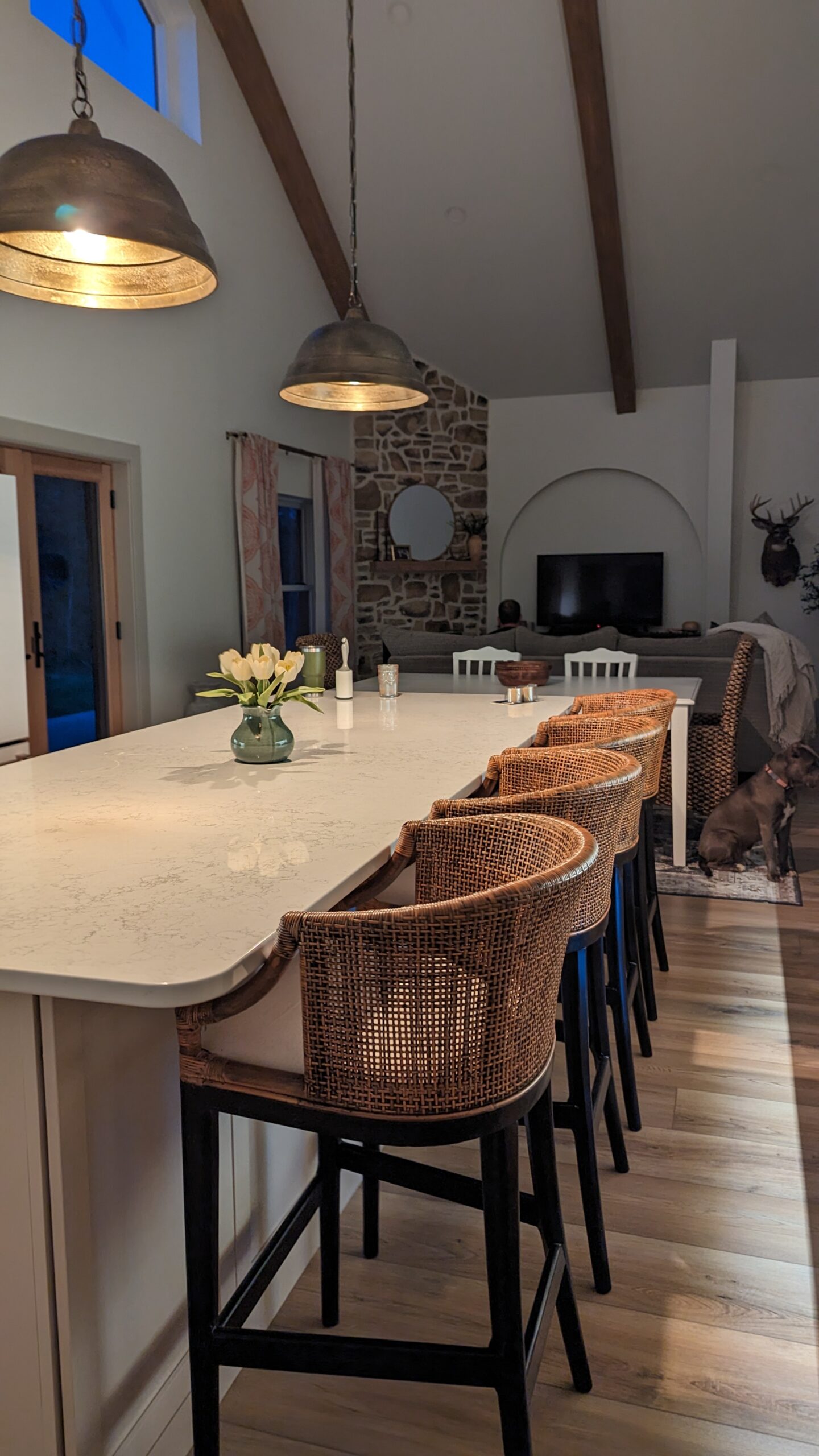 A cozy barndominium kitchen features a long marble island with wicker chairs, hanging pendant lights, and a vase of flowers. The background includes a stone fireplace, a TV, and birch tree wood beam ceiling accents. Warm and inviting atmosphere.