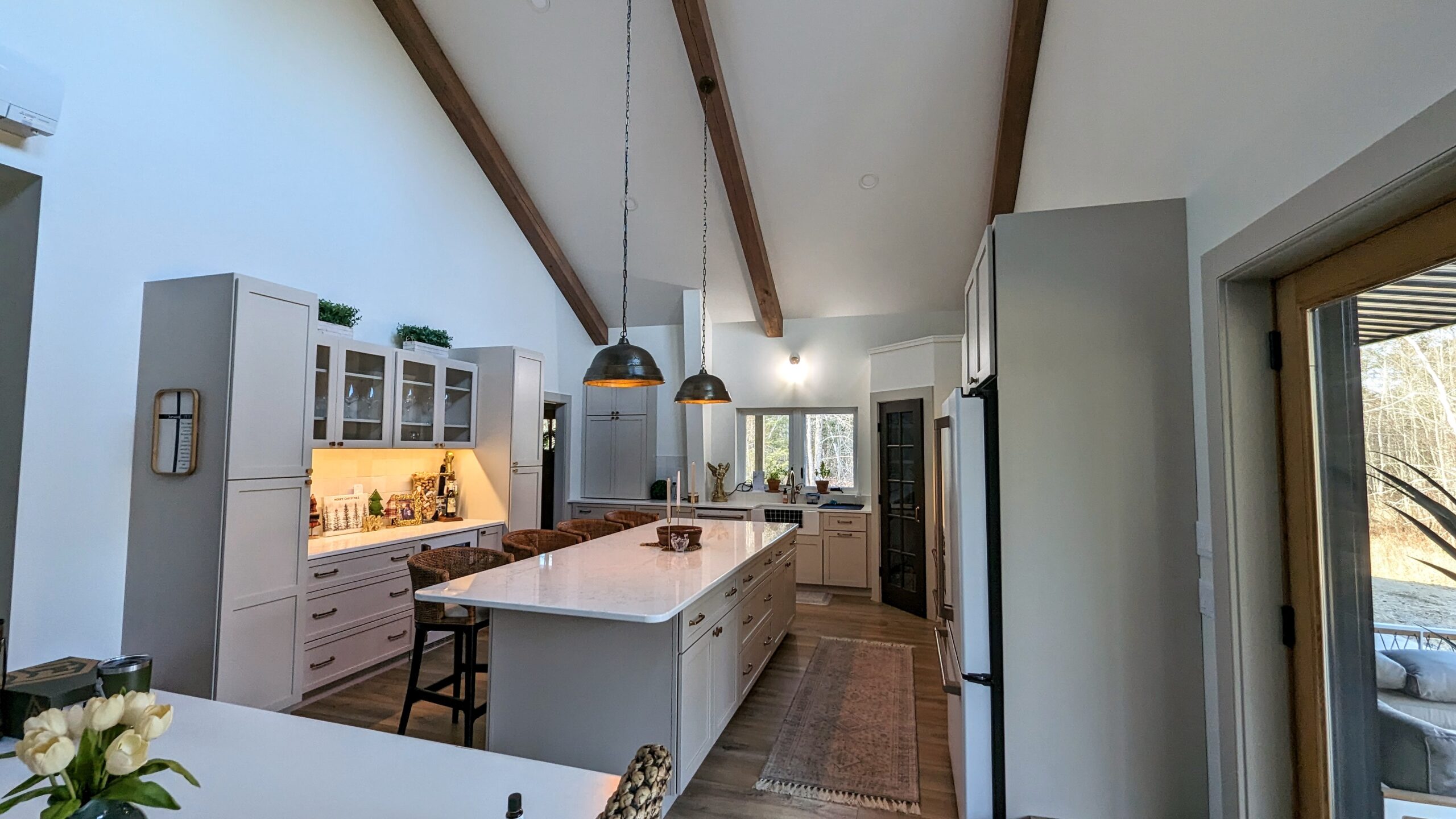 A spacious barndominium kitchen with high ceilings and exposed wooden beams features a large island with bar stools, pendant lights, modern appliances, and light-colored cabinets. A large window provides a picturesque view of the outdoor birch trees.