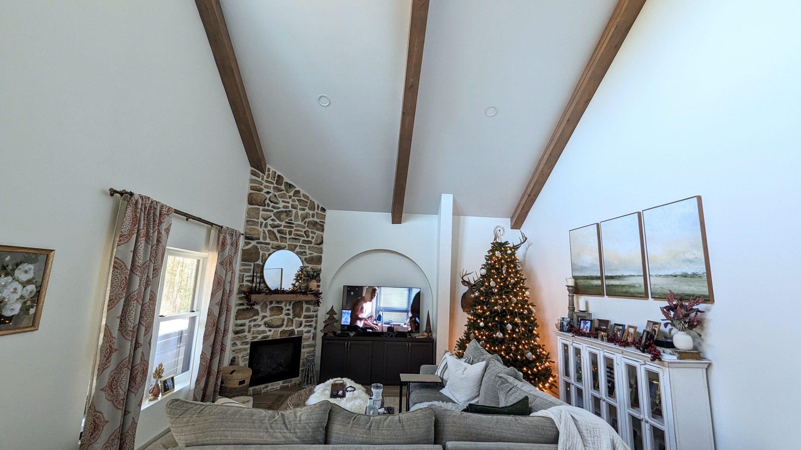 A cozy living room in a barndominium features high ceilings and wooden beams. Beside the stone fireplace, a Christmas tree with lights stands proudly. The sofa faces the TV, surrounded by light walls and decorated shelves, while large windows offer views of birch trees swaying outside.