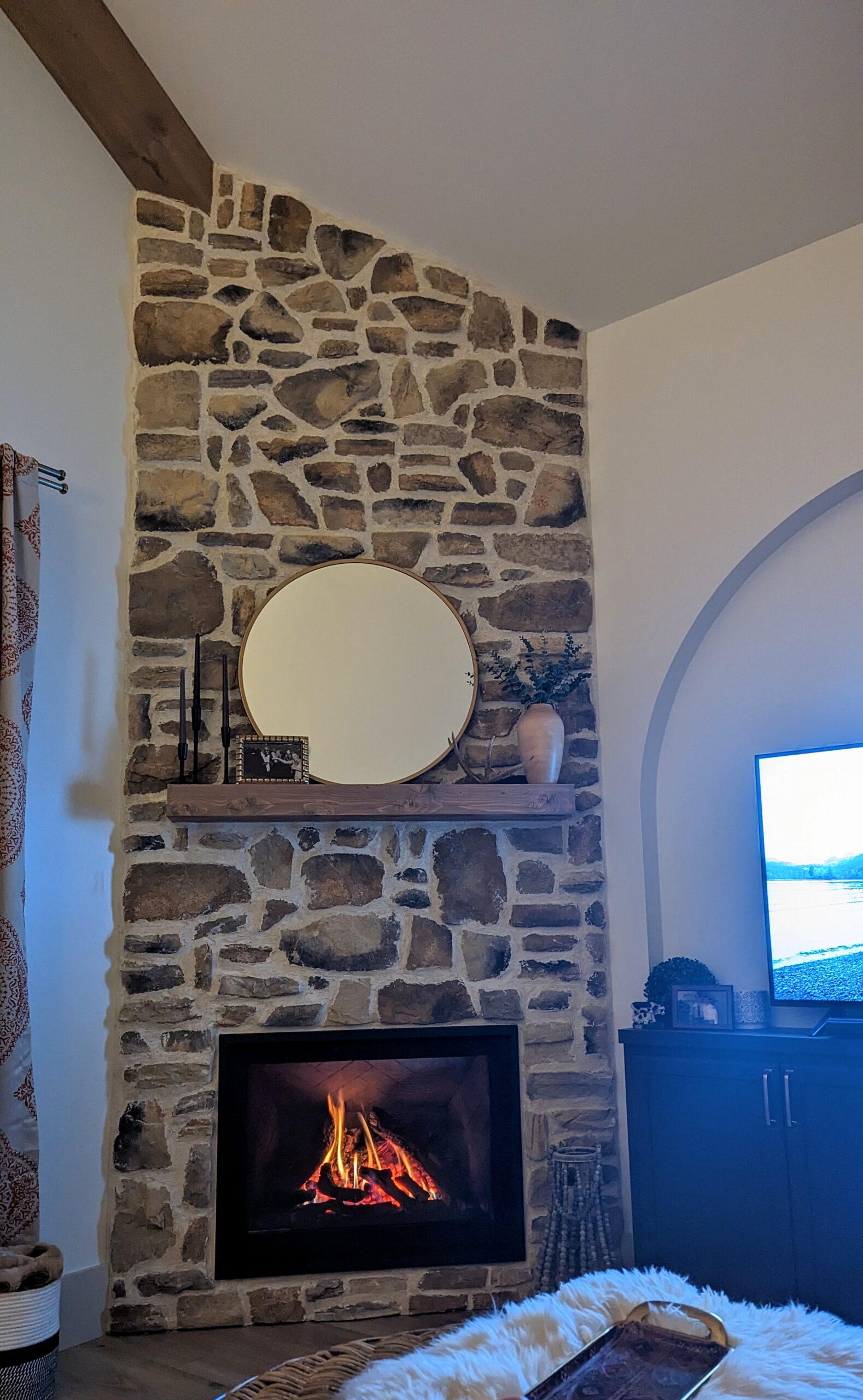 A cozy living room in a charming barndominium features a stone fireplace with a lively fire. Above it, on the wooden mantel, adorned with candles and a small plant, hangs a round mirror. A TV rests to the right, and a soft rug lies fronted by birch tree accents for added warmth.