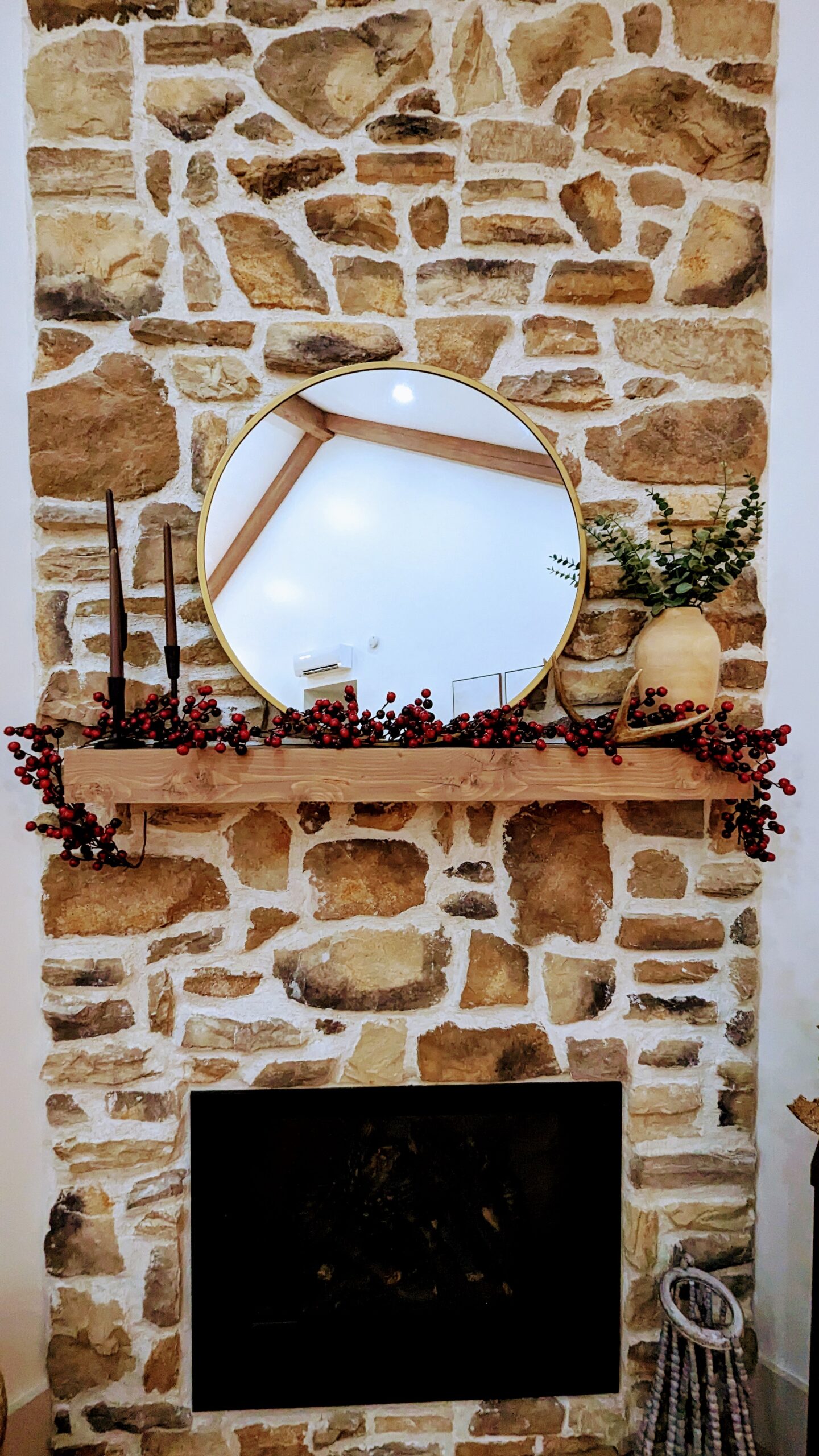 A rustic stone fireplace with a wooden mantel, reminiscent of a cozy barndominium, is adorned with red berries, a round mirror, and a vase filled with greenery. Birch tree accents join candles in holders on one side of the mantel.