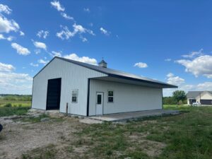 A large white metal barn with a grey roof stands on a concrete foundation in a grassy field. The barn has a tall sliding door on one side and a regular door with small windows on the other. The sky is blue with scattered white clouds.