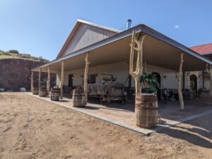 A rustic, covered patio area with wooden barrels and assorted vintage farming equipment is depicted. The structure has a corrugated metal roof supported by wooden posts. The ground around it is gravel with a dirt path. In the background, hills and clear blue sky are visible.