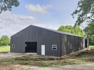 A large, dark gray metal barn with a gable roof stands amidst a rural, grassy area with a few trees in the background. The barn features several small windows, a couple of doors, and large openings for equipment or livestock access. The sky is partly cloudy.