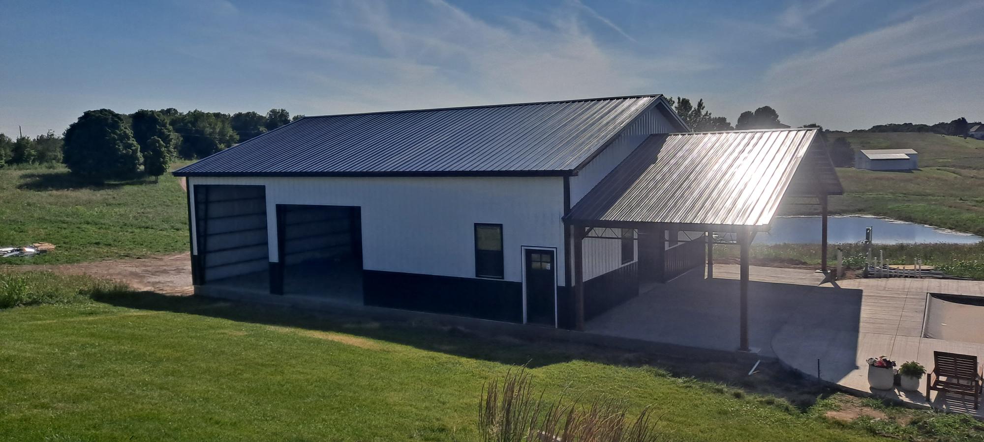 A large, modern barn with a white and black exterior and a shiny metal roof, standing on a grassy landscape. The barn features large sliding doors and an attached covered area with a concrete floor. A small pond and scattered trees are visible in the background.