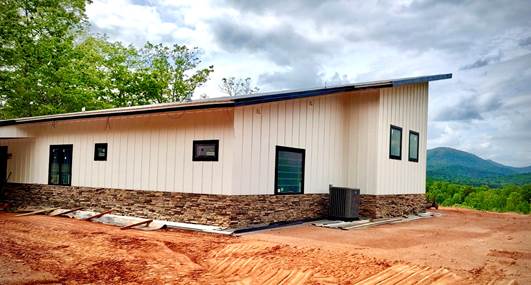 A modern single-story house with a sloped roof sits on a red dirt plot. It has white vertical panel siding and a stone foundation. There are several windows, and part of the structure is unfinished. Trees and mountains are visible in the background under a cloudy sky.