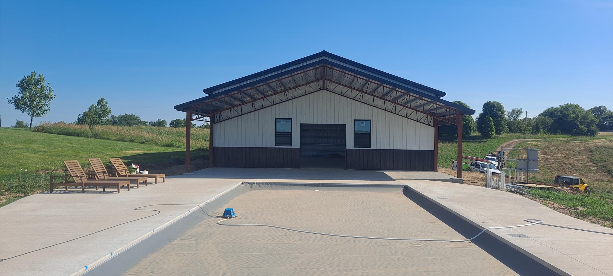 A modern barn-style building with a metal roof is situated in a rural area. In front of the building, there is an in-ground swimming pool under construction. To the left, several wooden lounge chairs are arranged on a concrete deck. The sky is clear and sunny.