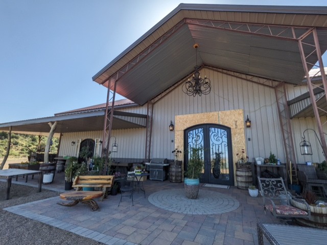 Image of a modern barn-style building with large double doors and a covered entryway. The patio area is furnished with wooden benches, potted plants, and rustic decor such as wine barrels and lanterns. The building is surrounded by a paved walkway.