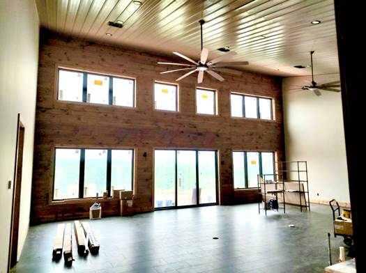 A spacious, unfinished room with high, wooden ceilings and a large wall of windows. Two ceiling fans hang from the ceiling. The room features large floor tiles and wooden beams on the floor. Shelving units and boxes are scattered around the room.