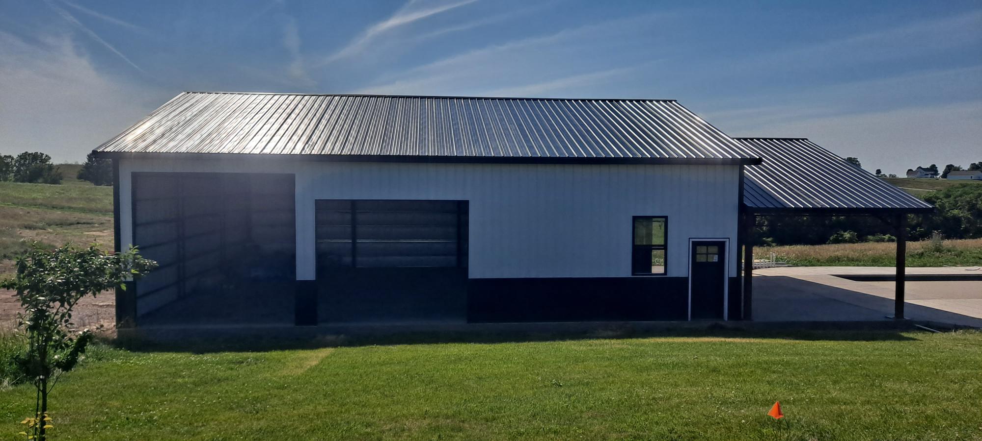 A large metal building with a black and white exterior, featuring two garage doors and a small entry door with a window. An attached carport extends from the right side. A grassy lawn is in the foreground, and the sky above is clear and blue.