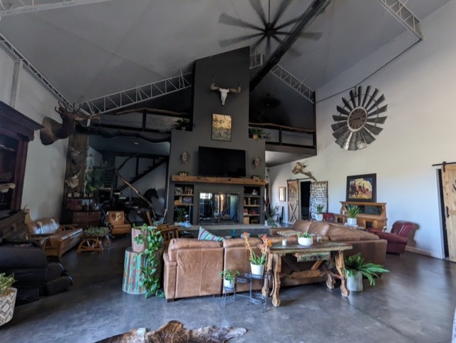 A spacious living room with high ceilings, featuring a large sectional sofa, rustic wooden furniture, and several mounted animal heads. A fireplace with a TV above it is centered on the far wall. Potted plants and decorative items are scattered throughout the room.