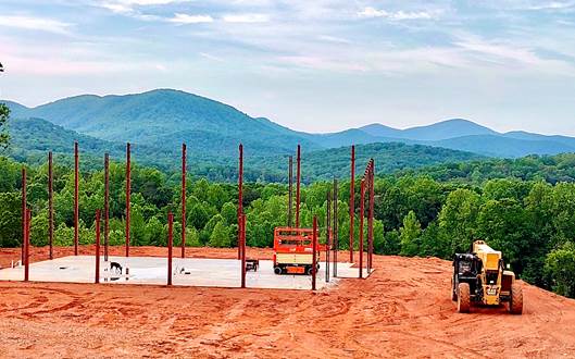 A construction site on red dirt, set against a backdrop of lush green hills and mountains. Several steel beams are erected on a rectangular concrete foundation, and construction vehicles, including a forklift, are present on site. The sky is light blue with scattered clouds.