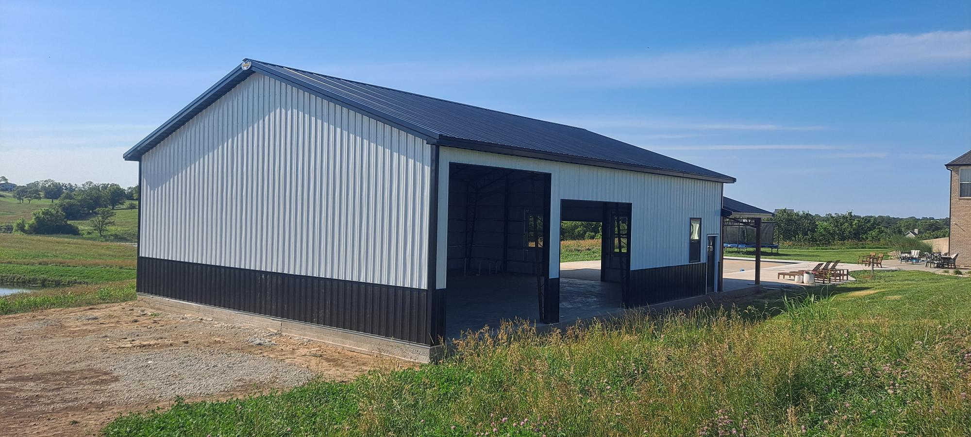 A large gray and black metal building with an open entrance and several windows, situated in a grassy, rural area under a clear blue sky. A small pond and more landscape are visible in the background.