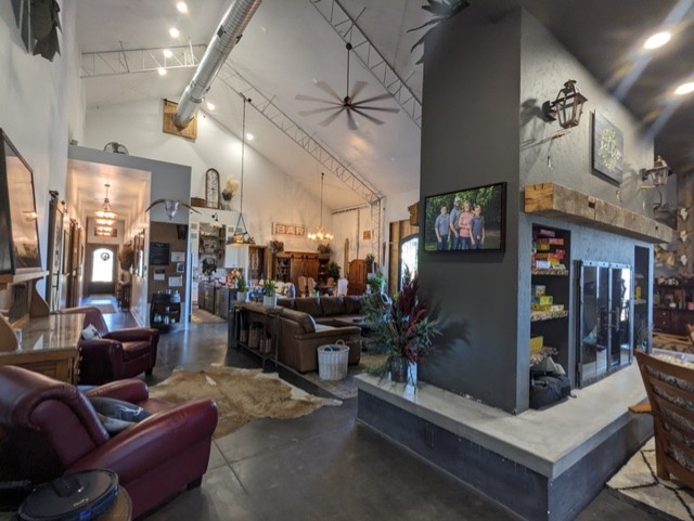 Spacious, rustic living area with high, angled ceilings adorned with ceiling fans. The space includes various seating areas, a fireplace with a TV mounted above, and a mix of modern and vintage decor. The room has polished concrete floors and eclectic decorations.