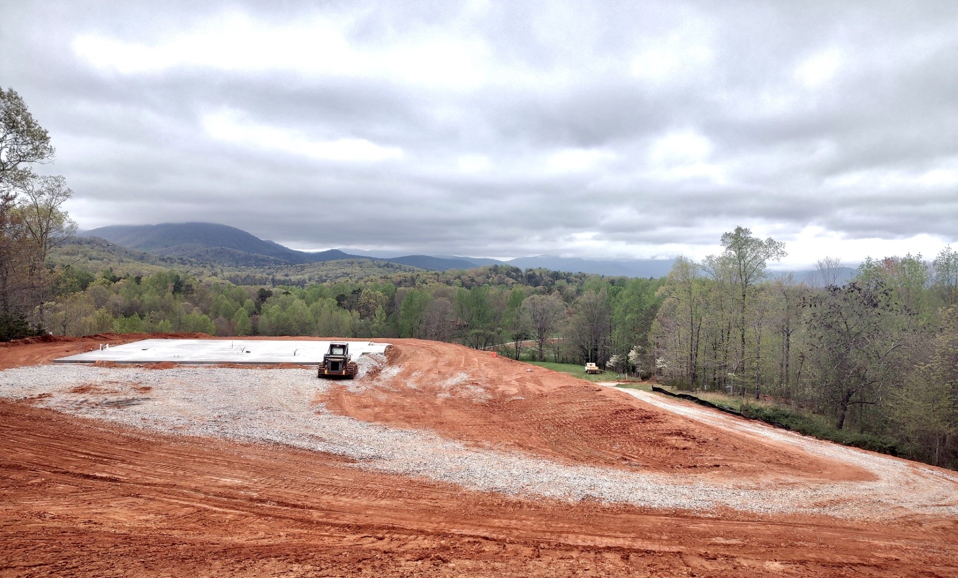 A construction site is shown with a concrete foundation in the foreground and a bulldozer working on the red dirt. Surrounding the site are grassy areas and trees, with green hills and a mountain range visible in the background under a cloudy sky.