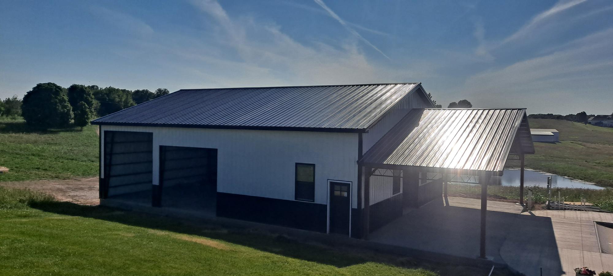 A large, modern metal building with a black roof is situated in a rural area. The structure includes a covered outdoor area to the right. The sky is clear with a few wispy clouds, and the sun is shining brightly, casting reflections on the roof. Nearby are open fields and trees.