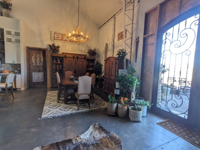 A well-decorated dining area with a rustic theme features a wooden table, cushioned chairs, a patterned rug, and a chandelier made of antlers. Potted plants, wooden cabinets, and a large decorative wrought iron door complete the cozy and inviting atmosphere.