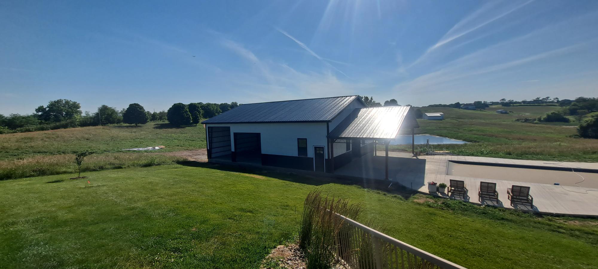 A large white and gray barn-style building with a metal roof stands in a grassy field under a clear blue sky. There is a small pond nearby and several lounge chairs on a paved section of the lawn. Rolling hills and trees are visible in the background.