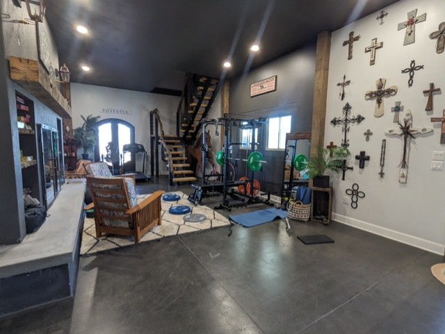 A room with a workout area featuring gym equipment like a squat rack and weights. There's a wooden staircase leading to an upper level, various crosses mounted on the wall, and a comfortable seating area with wooden chairs and a bench. The lighting is warm.